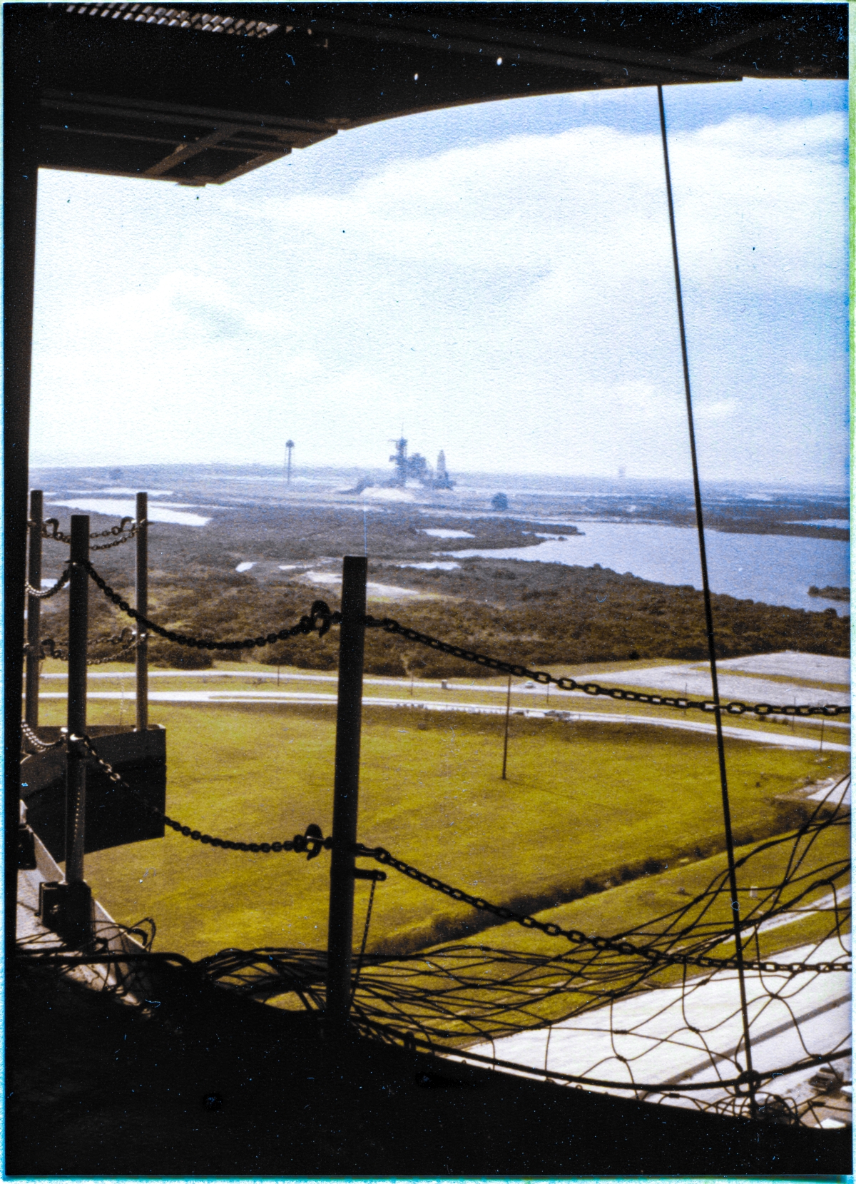 Image 040. Viewed from the platform decking at elevation 212’-2” inside the RCS Room on the Rotating Service Structure at Launch Complex 39-B, Kennedy Space Center, Florida, the Space Shuttle Columbia can be seen in the distance, carried by the Mobile Launch Platform, near the top of the Pad Slope at Pad 39-A, nearing the silhouette of the demated RSS on A Pad, completing its very slow rollout journey which started inside the VAB in predawn darkness. This is only the second time a Space Shuttle has been rolled to a launch pad, and when it flies, it will be the very first time ever that a Space Shuttle gets reused, thus validating the initial premise of reusable spacecraft which the Shuttle embodies. Your point of view on the RSS is in the area, when it is swung around and mated with a Space Shuttle, where the left side of the Space Shuttle’s nose, immediately forward of the Crew Cabin, will be occupying the empty space you now see, and if that were the case, none of the distant landscape would be visible, and instead you would be seeing the tile-covered nose of the Orbiter, in the area where the openings in the body of the Shuttle for the Forward Reaction Control System Thrusters are located. In the immediate foreground, removable handrails and safety chains, and some safety netting, can be seen guarding the sheer drop to the Pad Deck which yawns menacingly just ahead of you. Photograph by James MacLaren.